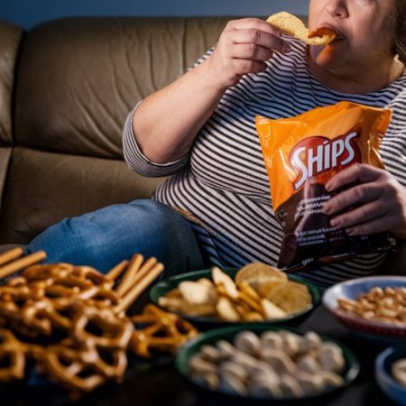 Žena s nadváhou na gauči večer u televize pojídá chipsy a je obklopená slanými pochutinami